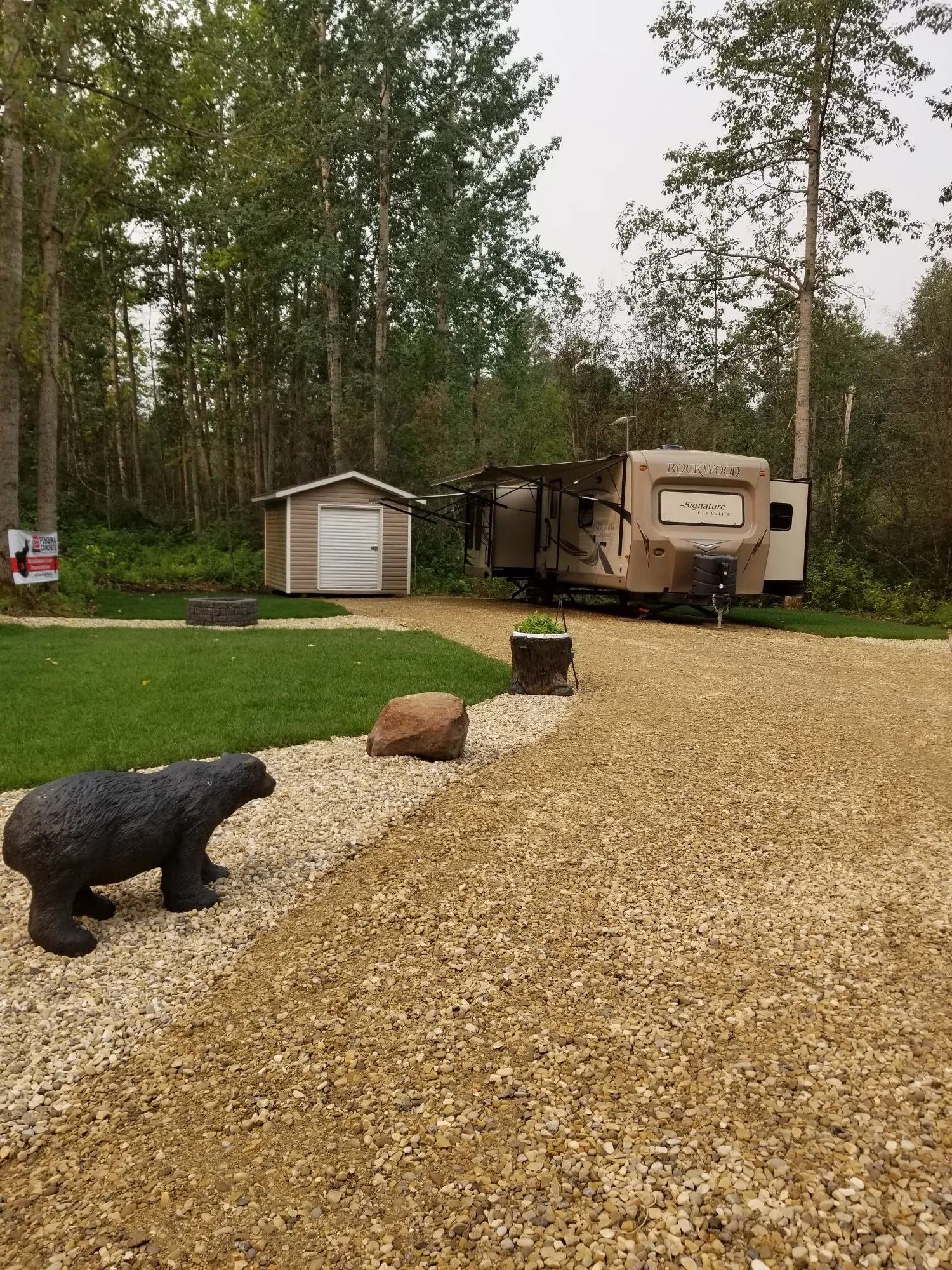 RV parked on a gravel pad near a small shed with decorative bear statue and surrounding trees.