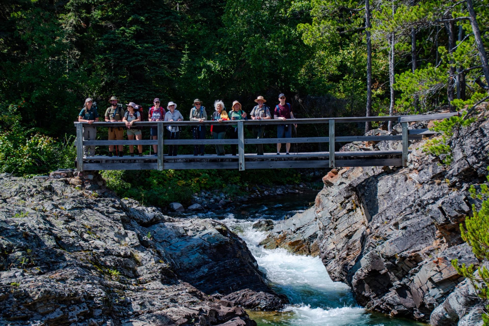 Guided hikers stand behind a wooden fence over a rocky stream in forest.