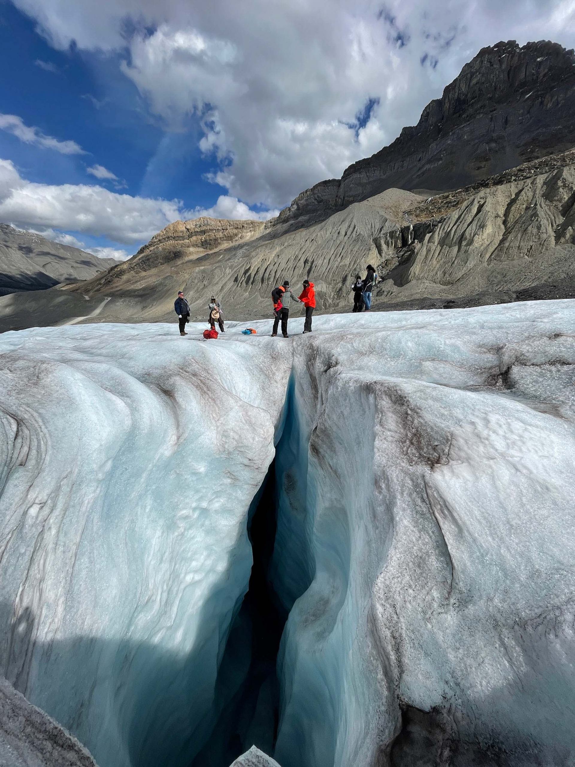 Wide glacier crevasse with people standing nearby, surrounded by rocky mountains and clouds.