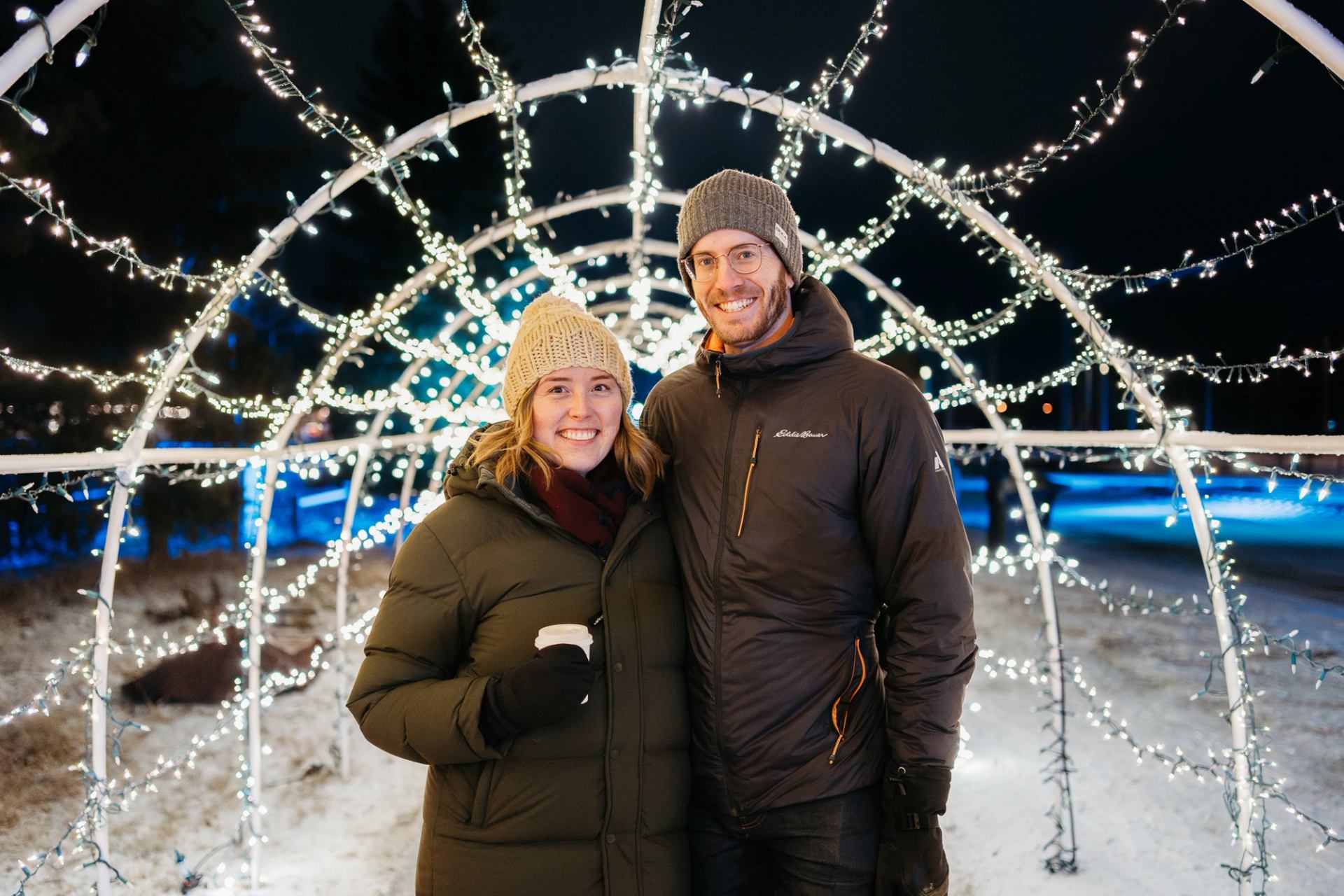 Two people standing under a dome of twinkling white holiday lights at an outdoor winter market.