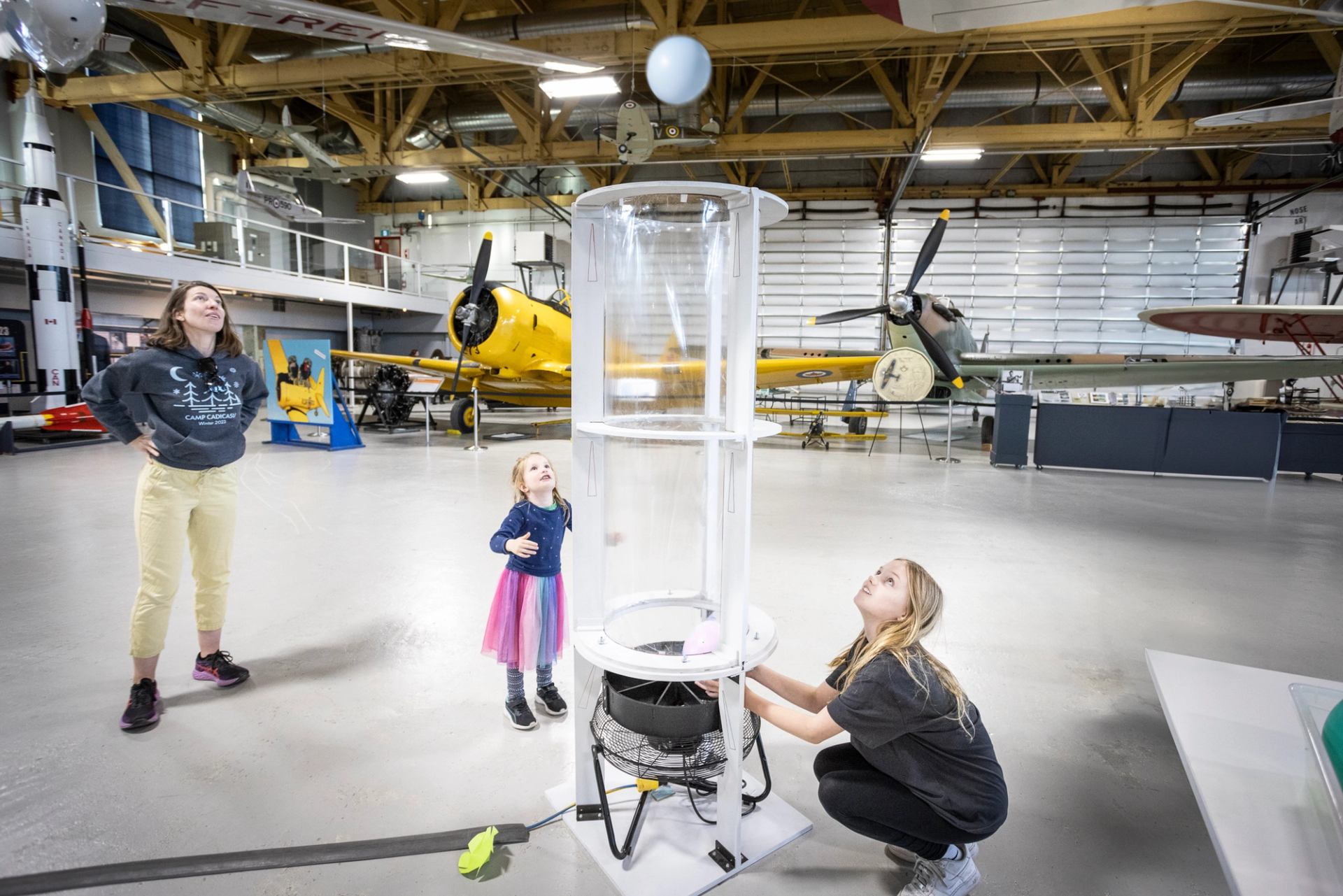 Visitors explore a wind tunnel exhibit with vintage planes in the background at Hangar Museum.