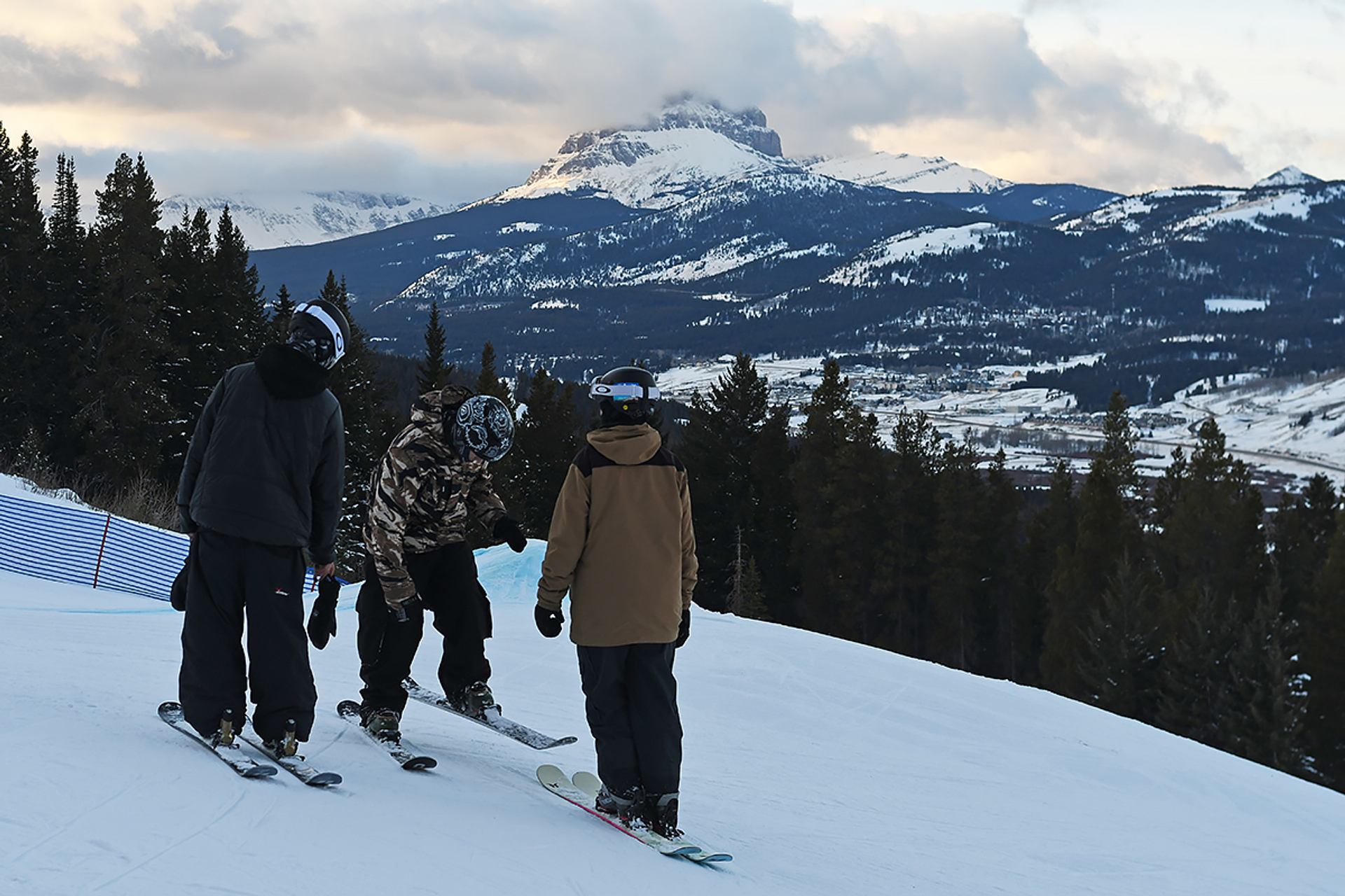 Three skiers standing on a snowy slope overlooking a mountain valley at sunset.