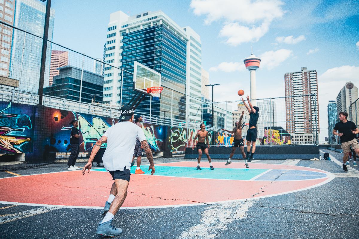 People play basketball on a colorful outdoor court, with tall buildings and Calgary tower behind them.