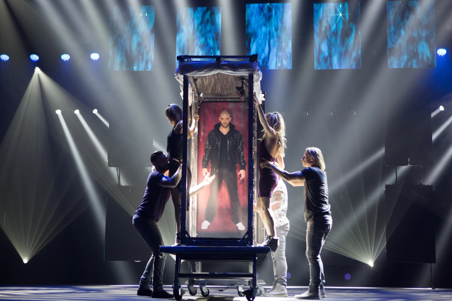 Stage illusionist stands inside tall glass box as assistants adjust frame under spotlights.