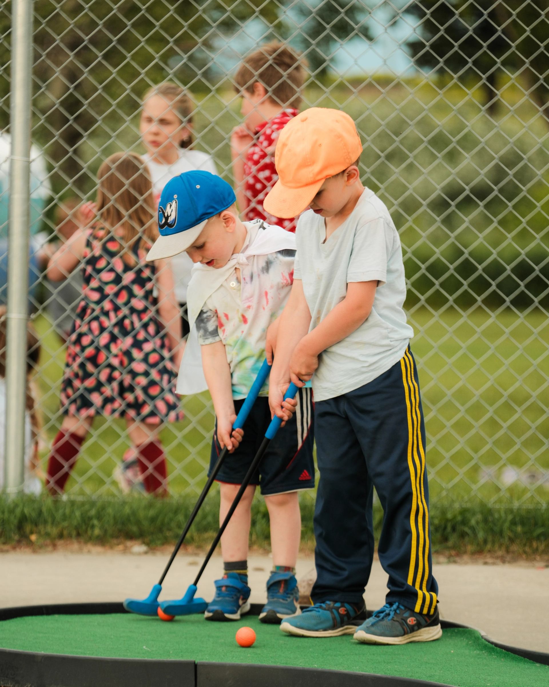 Two kids play mini-golf outdoors near a fence with greenery in the background.