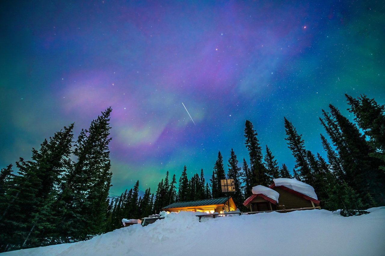 Snow-covered cabins at night with vivid green and purple aurora borealis filling the sky.