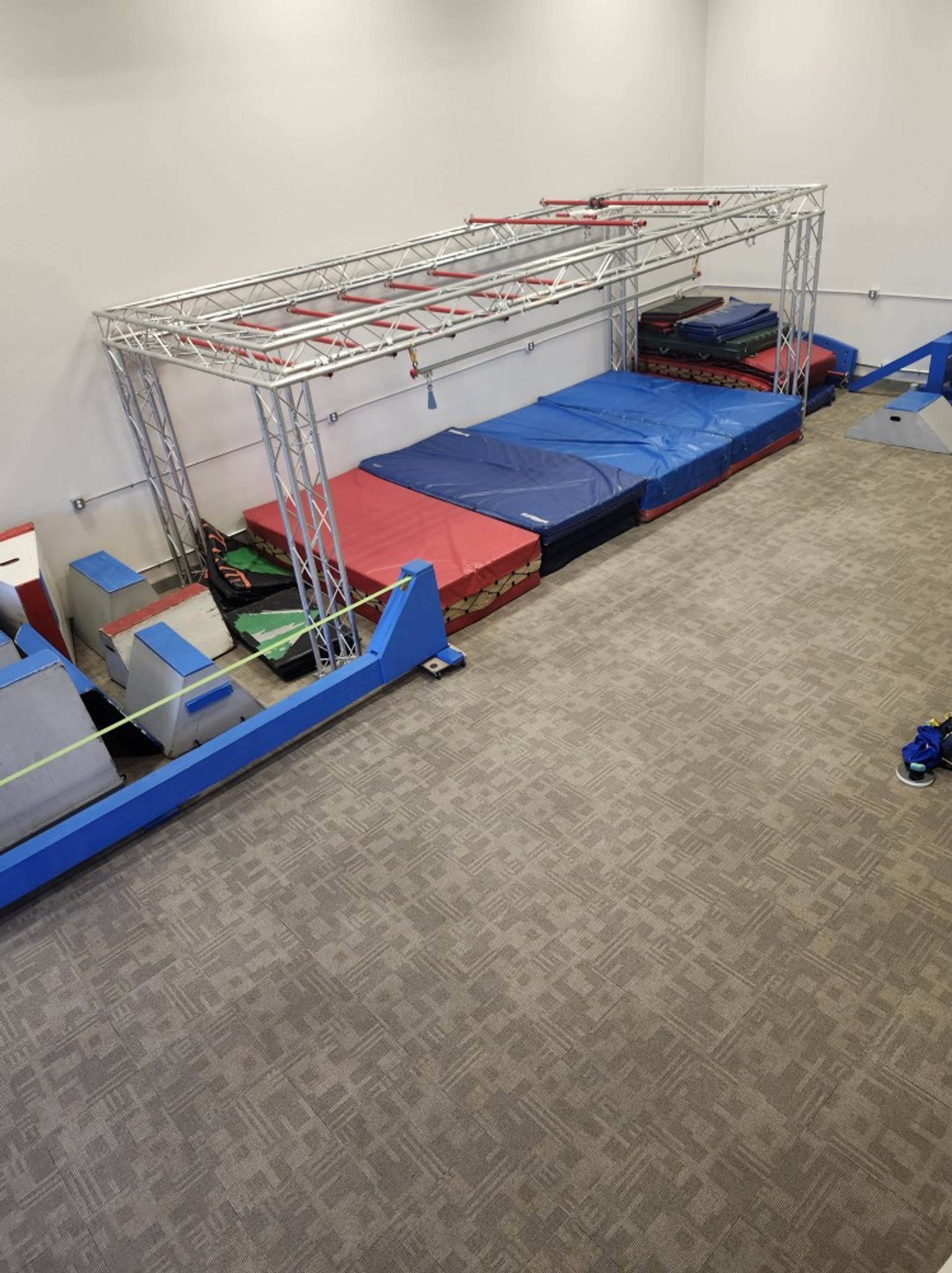 An indoor ninja warrior course with monkey bars, various obstacles, and safety mats on a carpeted floor.