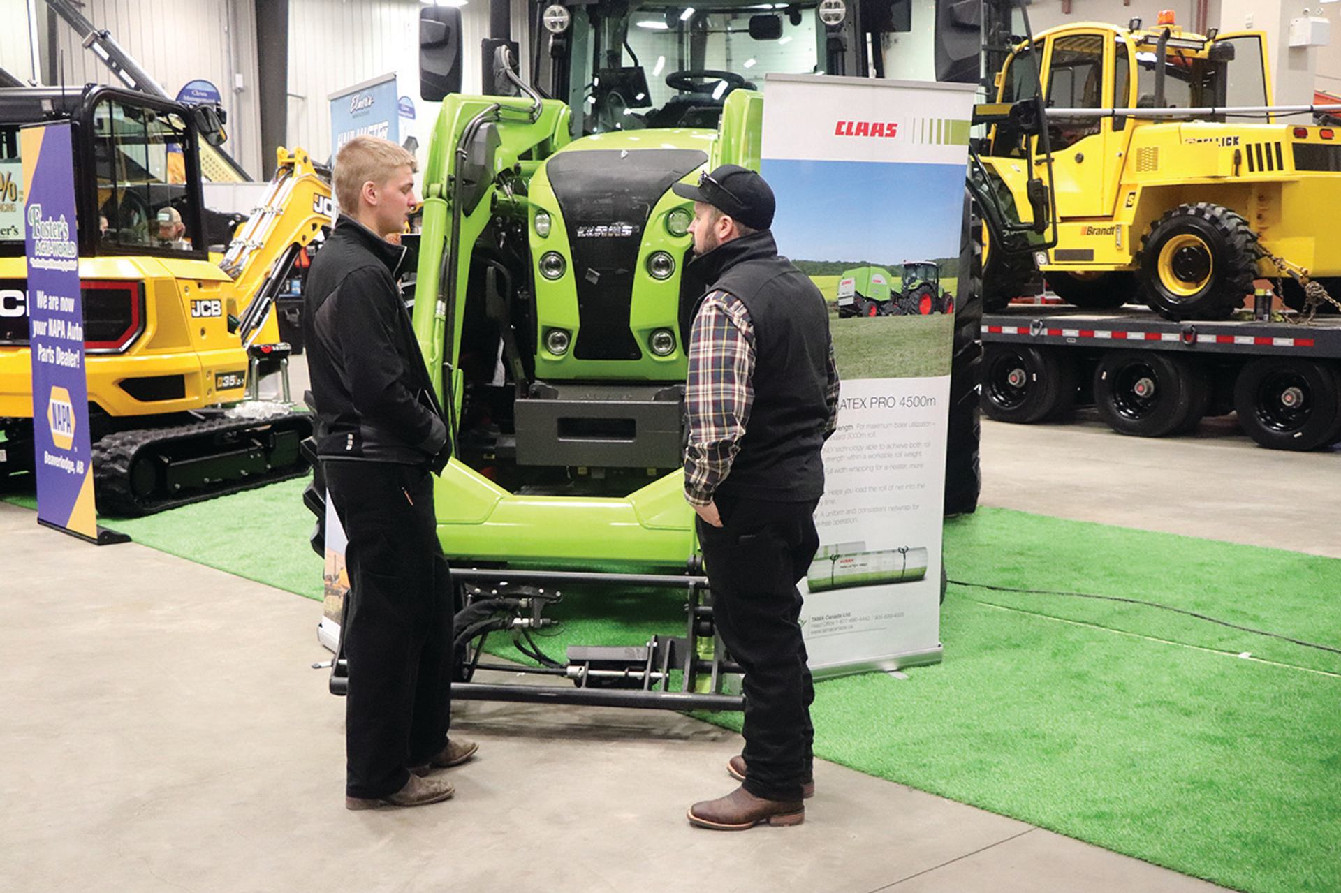 Two men stand and talk next to a bright green piece of agricultural machinery at an indoor trade show, with other yellow equipment in the background.