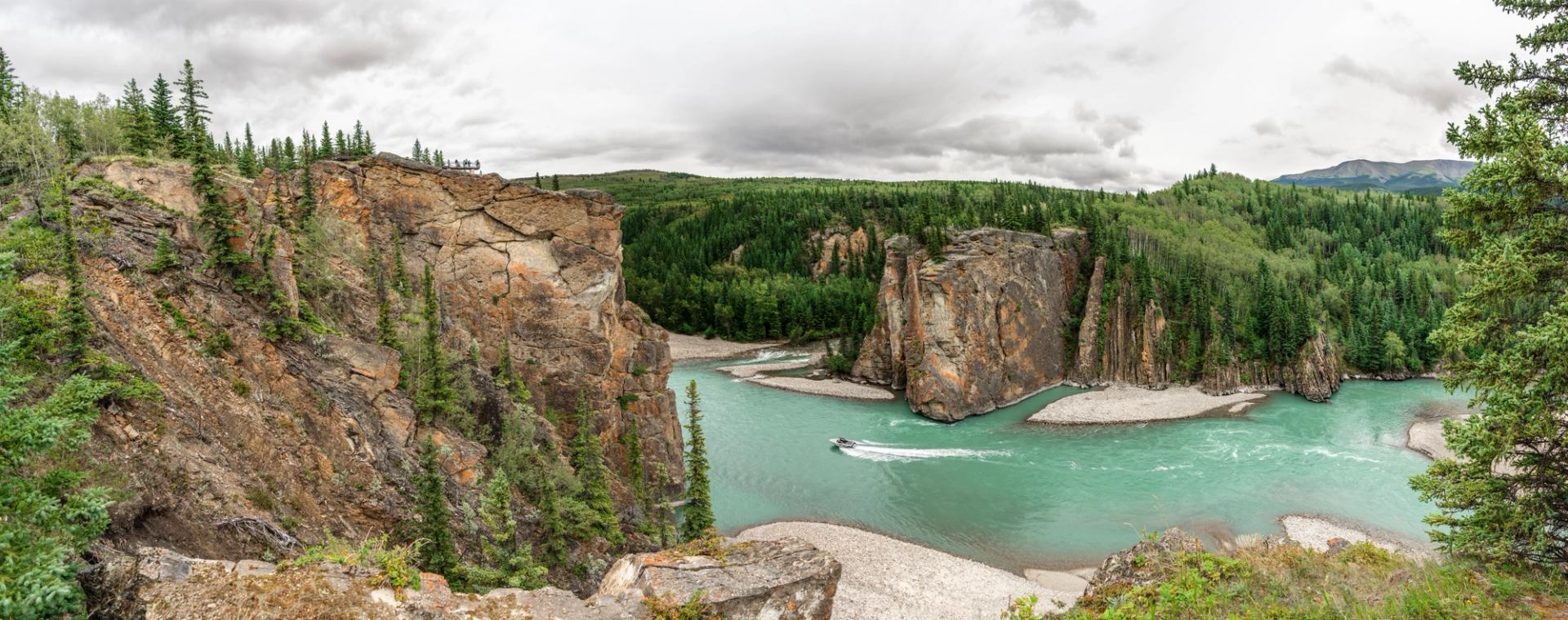 Turquoise river winds between rocky cliffs and evergreen forest beneath cloudy skies.