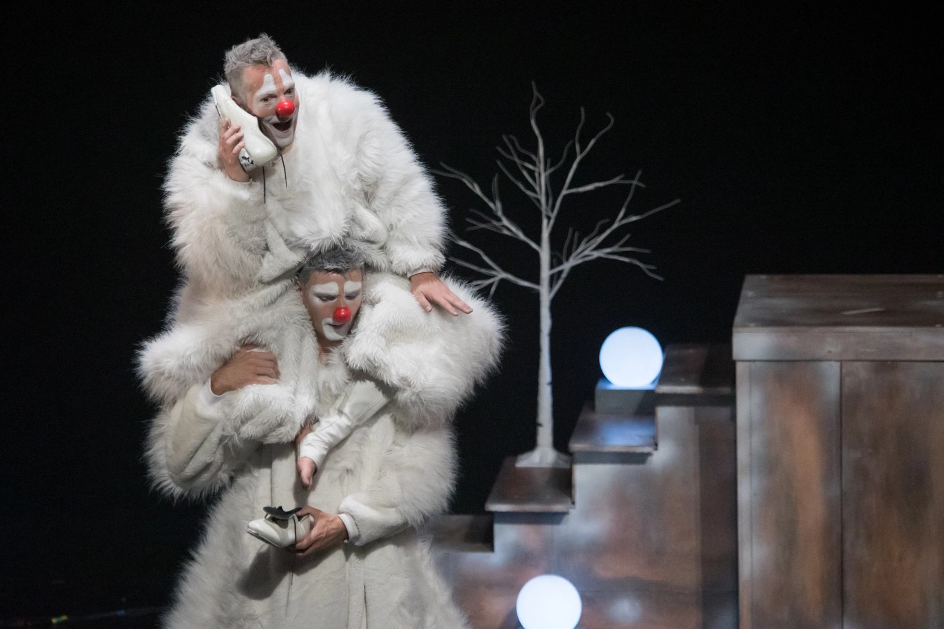 Two clowns in white fur costumes perform onstage beside bare tree and glowing orbs.