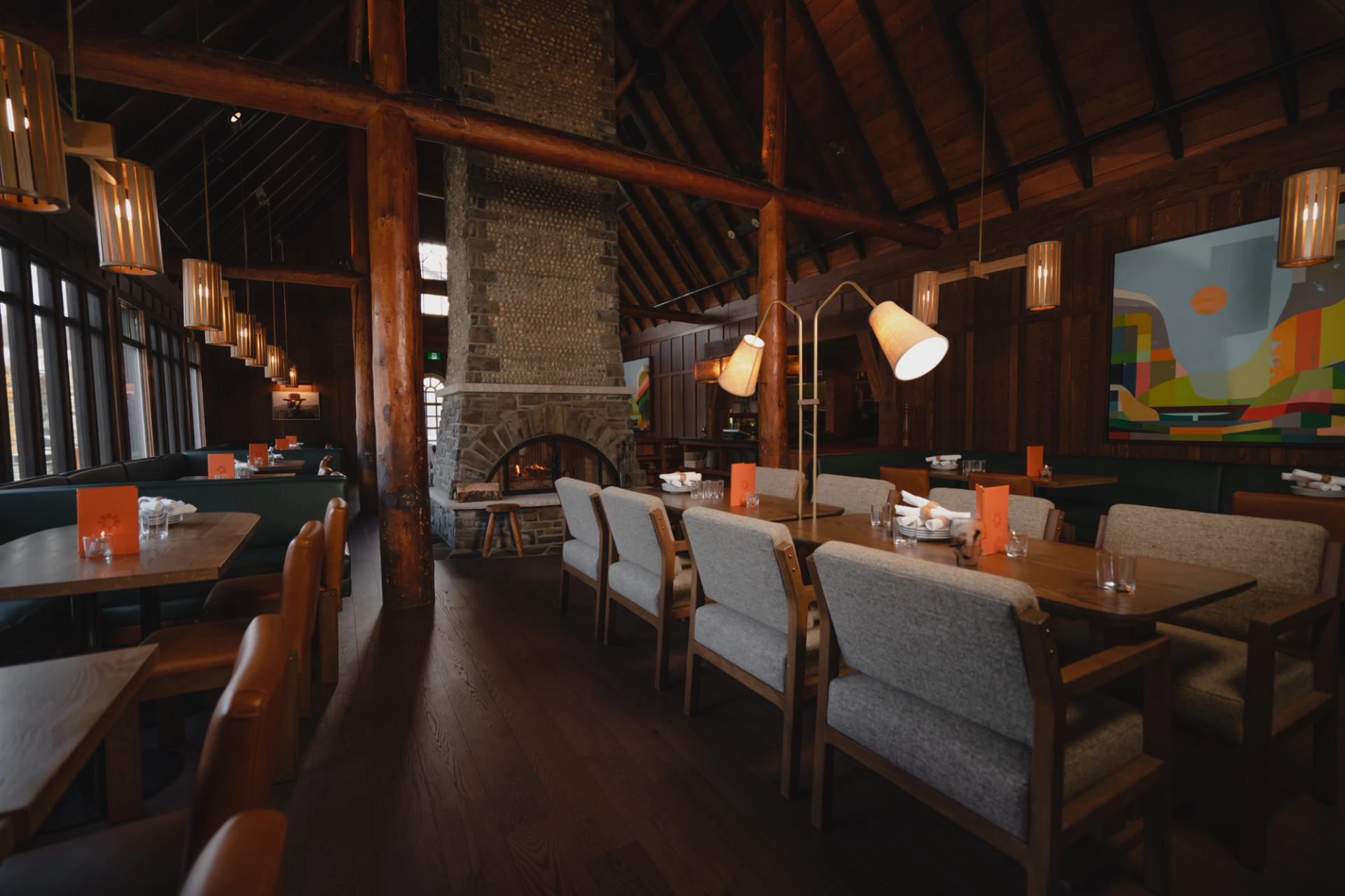 A rustic-modern restaurant dining room with wooden beams, a stone fireplace, and contemporary tables and chairs.