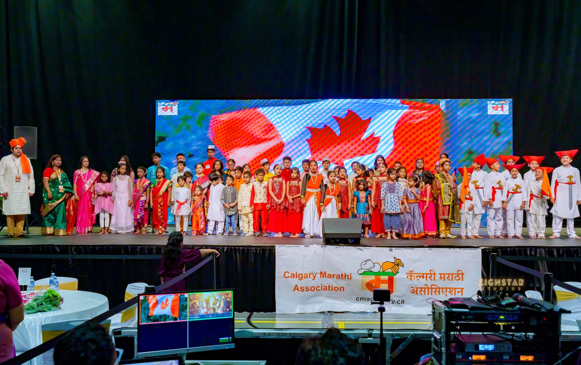 Children in colourful traditional clothing perform before a Canadian flag display.