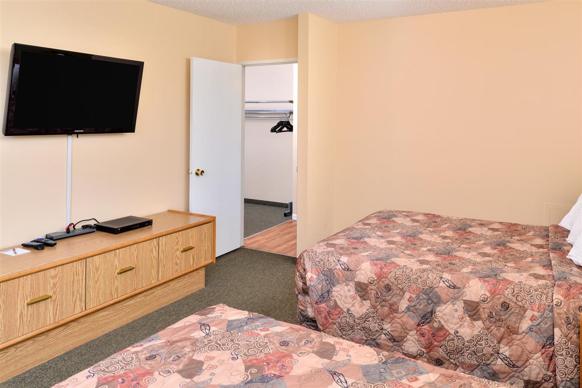 Guest room features two beds, a wall-mounted television, dresser, and open closet space nearby.