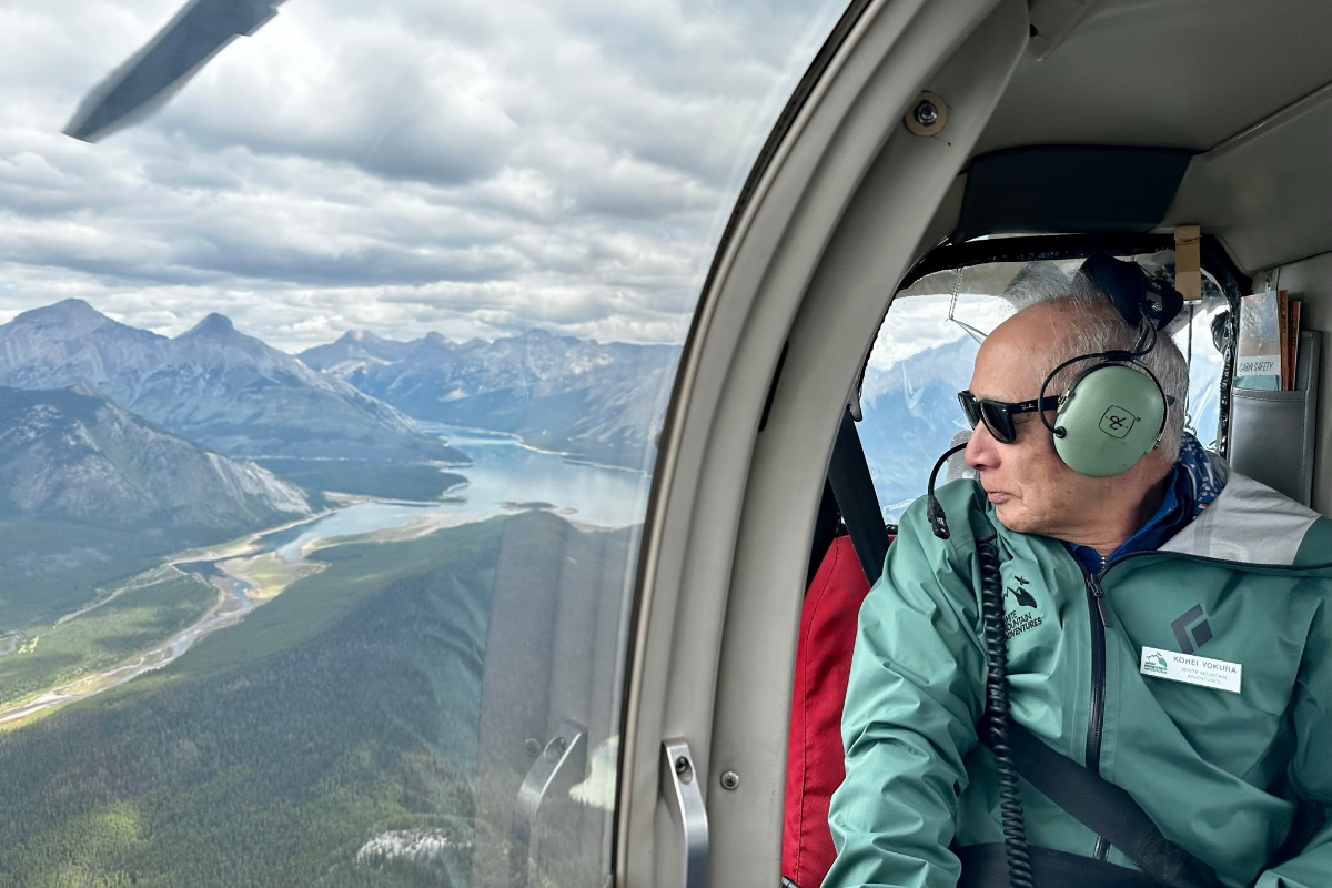 Helicopter passenger wearing headset looks out window at mountains, river and forest below.