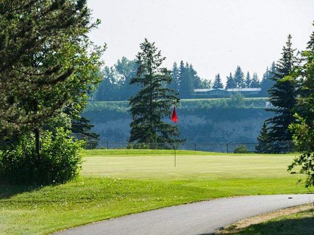 Golf green with red flag beside paved path, trees and distant ridge.