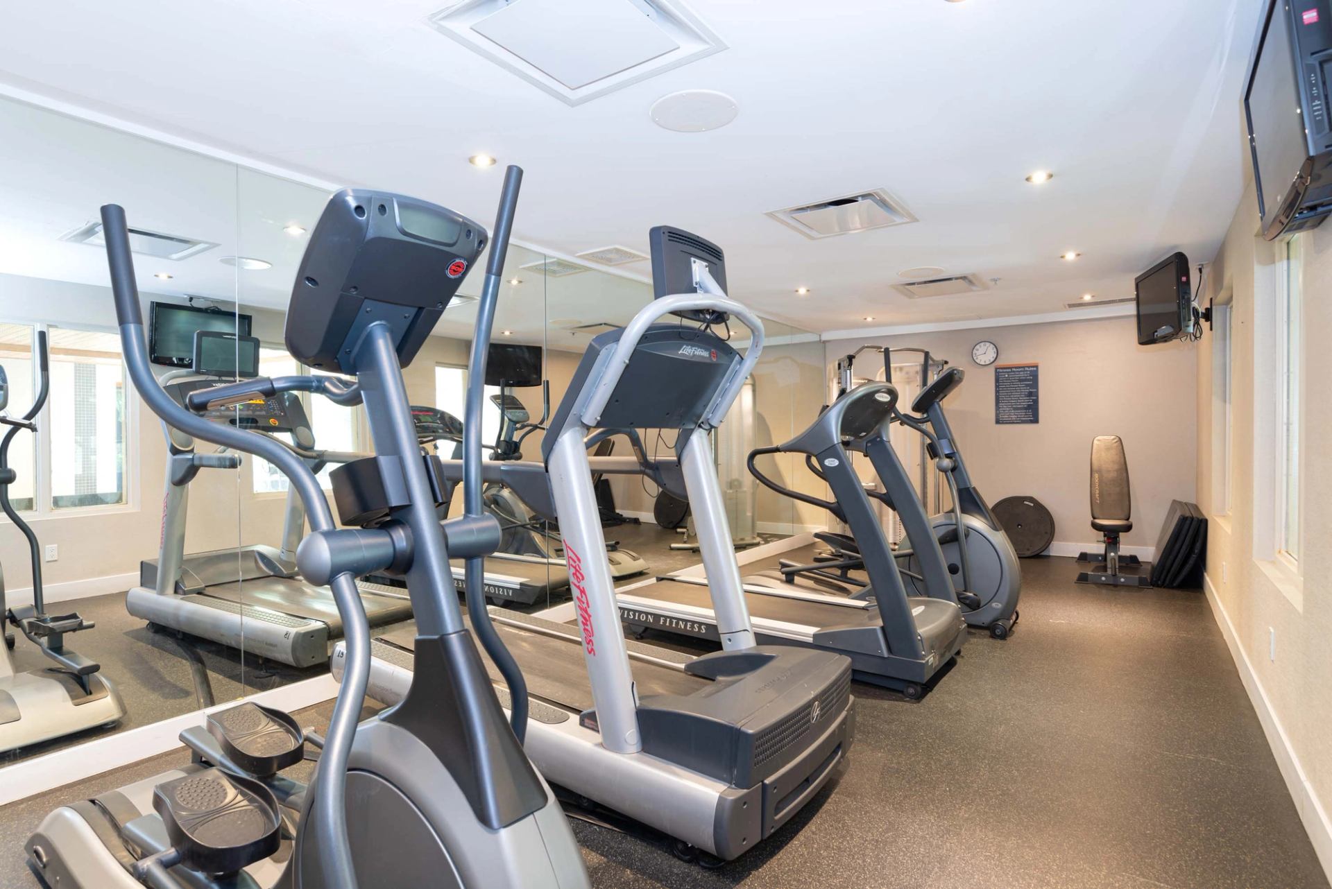 Small fitness room with treadmills, elliptical machines, mirrors and wall-mounted televisions.