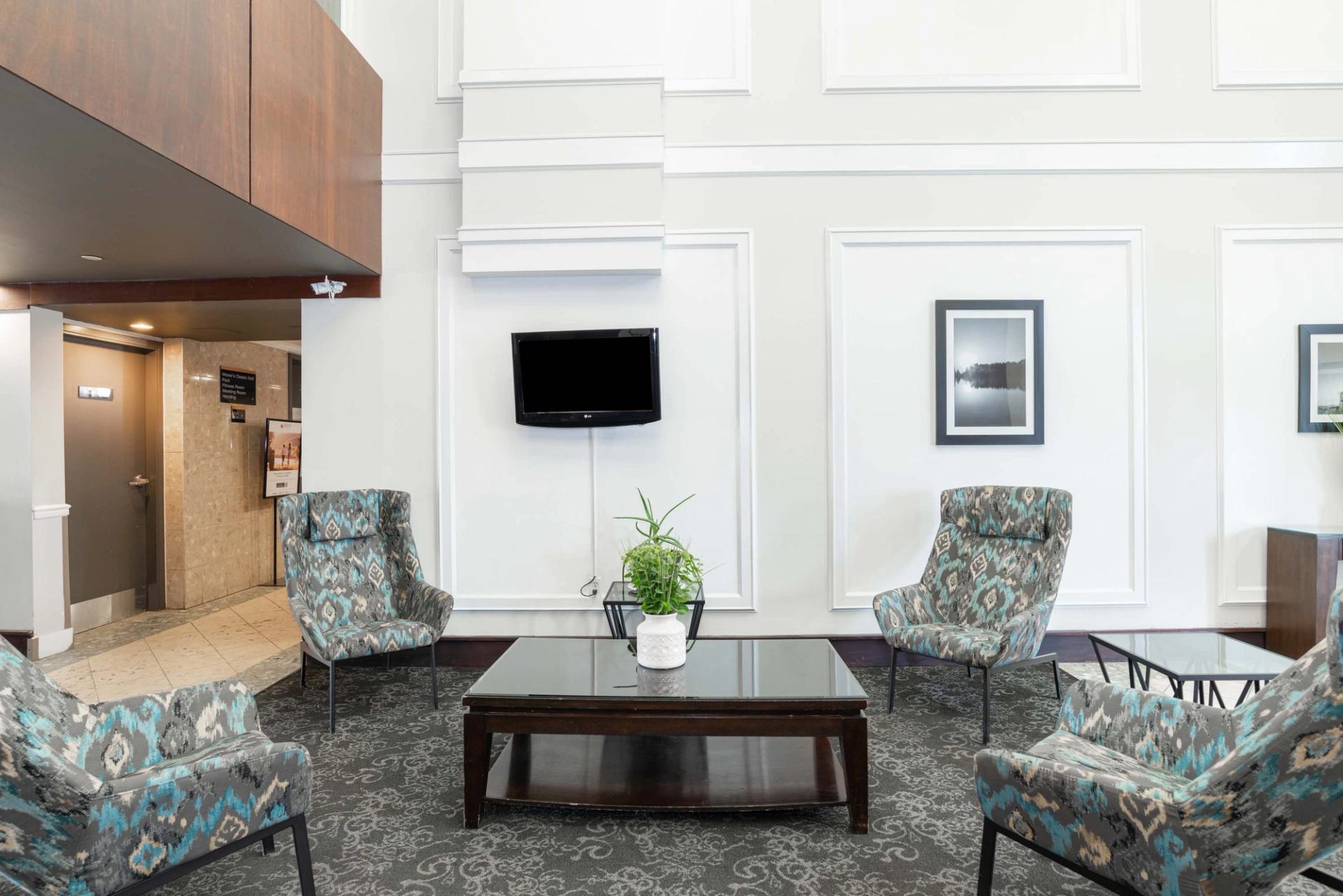 Lobby seating area with patterned chairs, glass coffee table, wall-mounted TV and artwork.