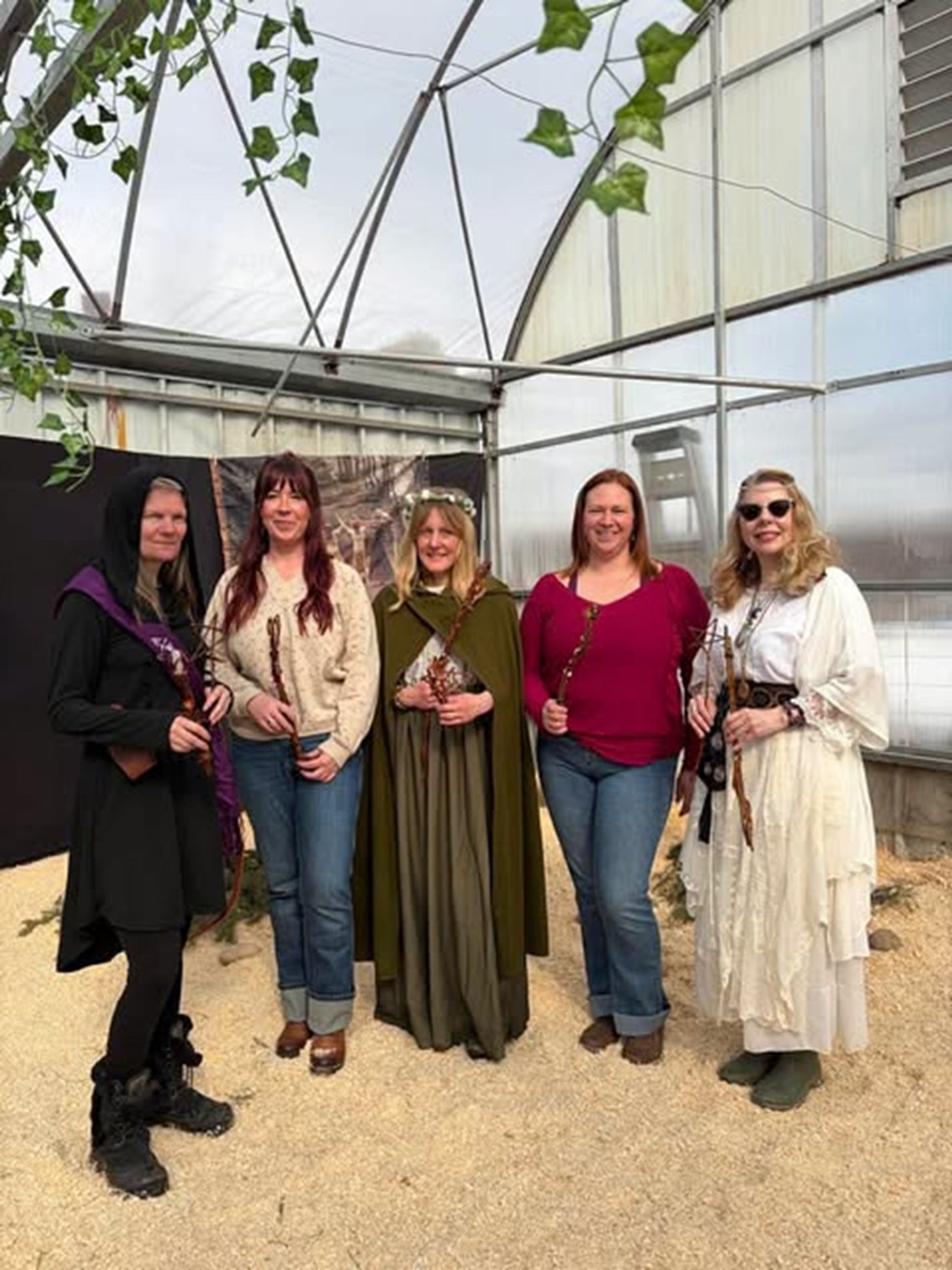Five visitors pose inside a greenhouse wearing medieval-inspired clothing and holding wooden staffs.