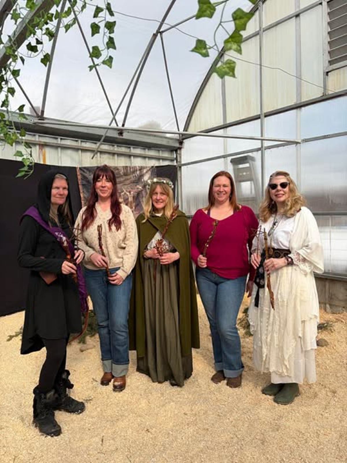 Five visitors pose inside a greenhouse wearing medieval-inspired clothing and holding wooden staffs.