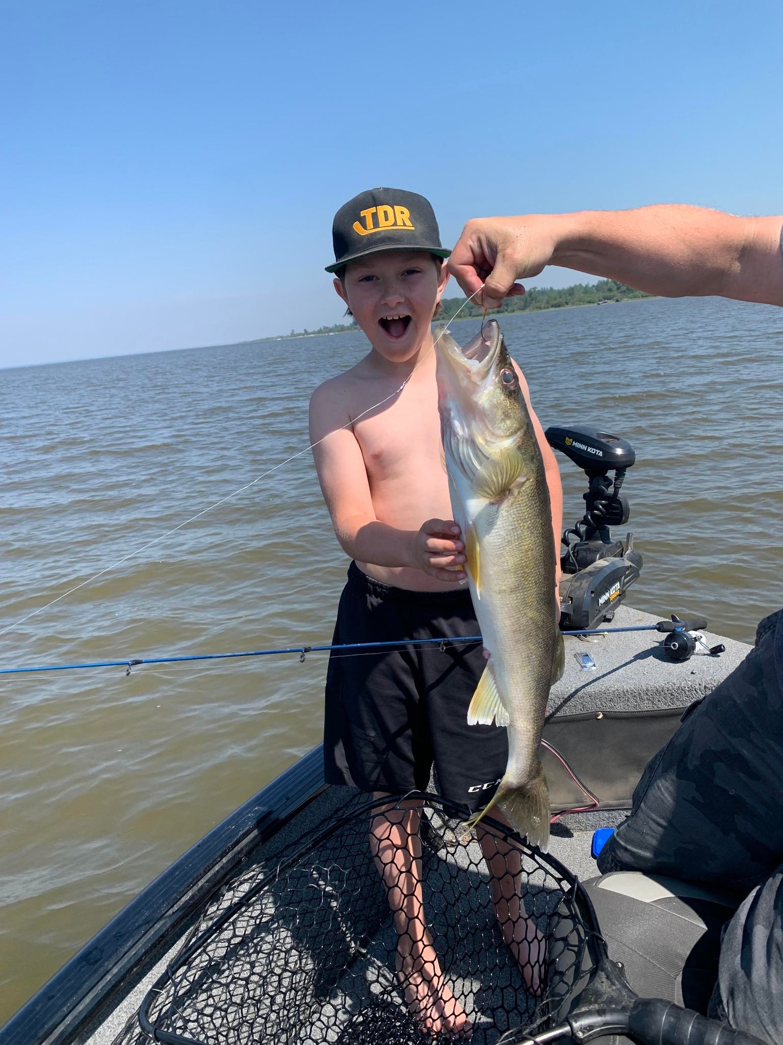 Shirtless child stands in a fishing boat beside a large fish held by an adult.