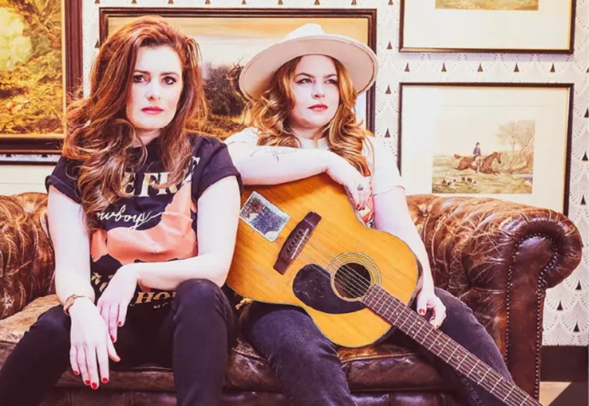 Two people sit on a leather couch, one holding an acoustic guitar.