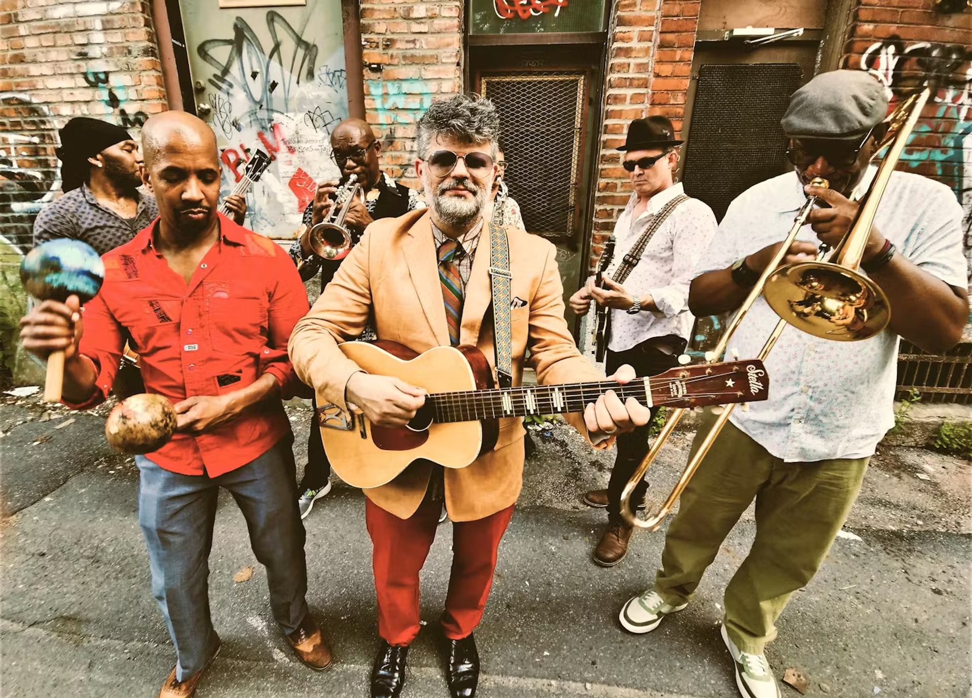Kobo Town performs in a graffiti-lined alley with brass, guitar, and maracas.