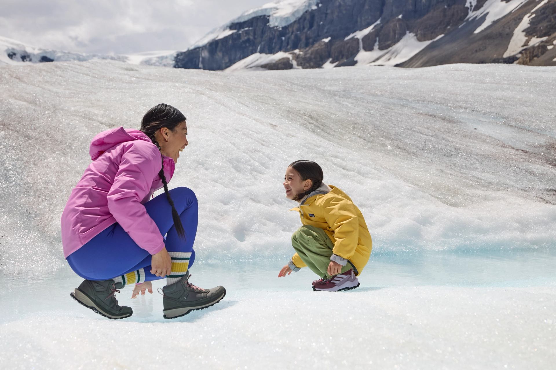 Two people in bright jackets crouch on a snowy glacier near blue ice.