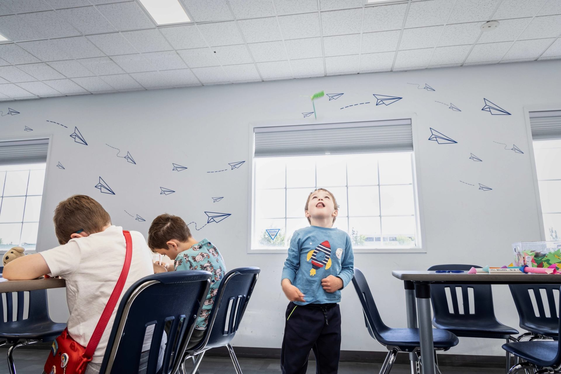 Children sit and stand among classroom tables beneath paper airplane wall drawings.