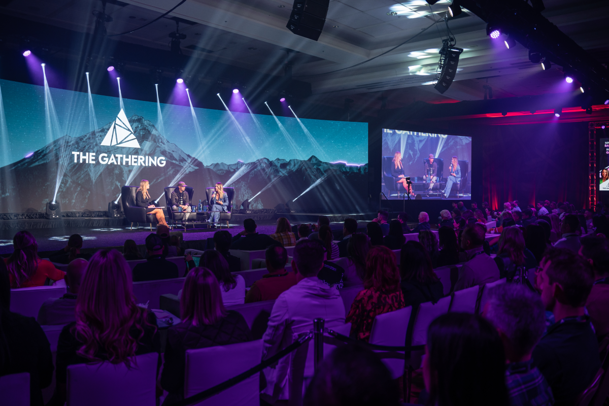 Live conference stage with panelists, large screens, and audience under purple spotlights.