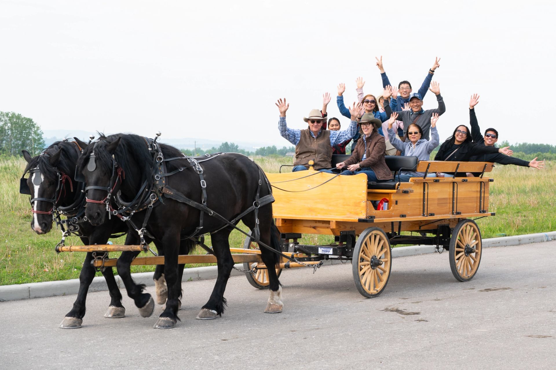 Group rides horse drawn wagon and waves while traveling along road at Stampede event.