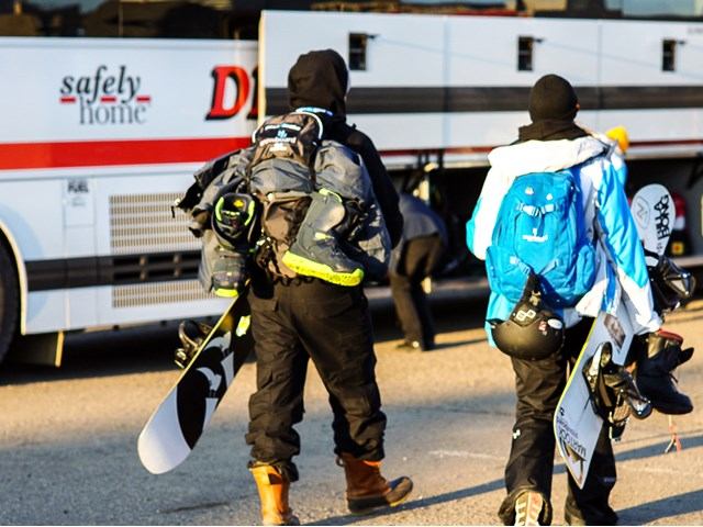 Snowboarders in winter gear walk toward Diversified bus with “safely home” text on the side.