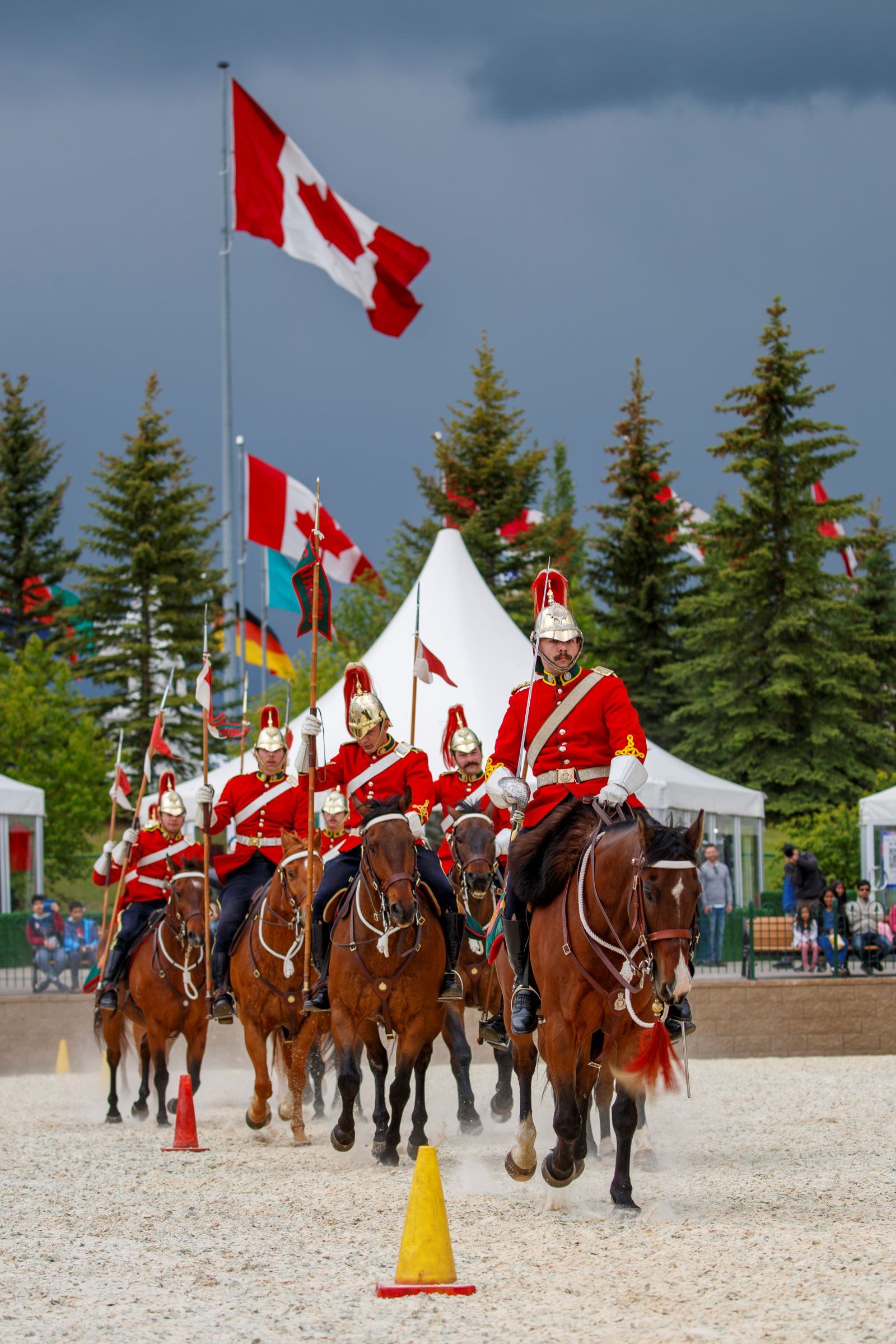A group of uniformed riders on horseback perform a parade beneath Canadian flags.