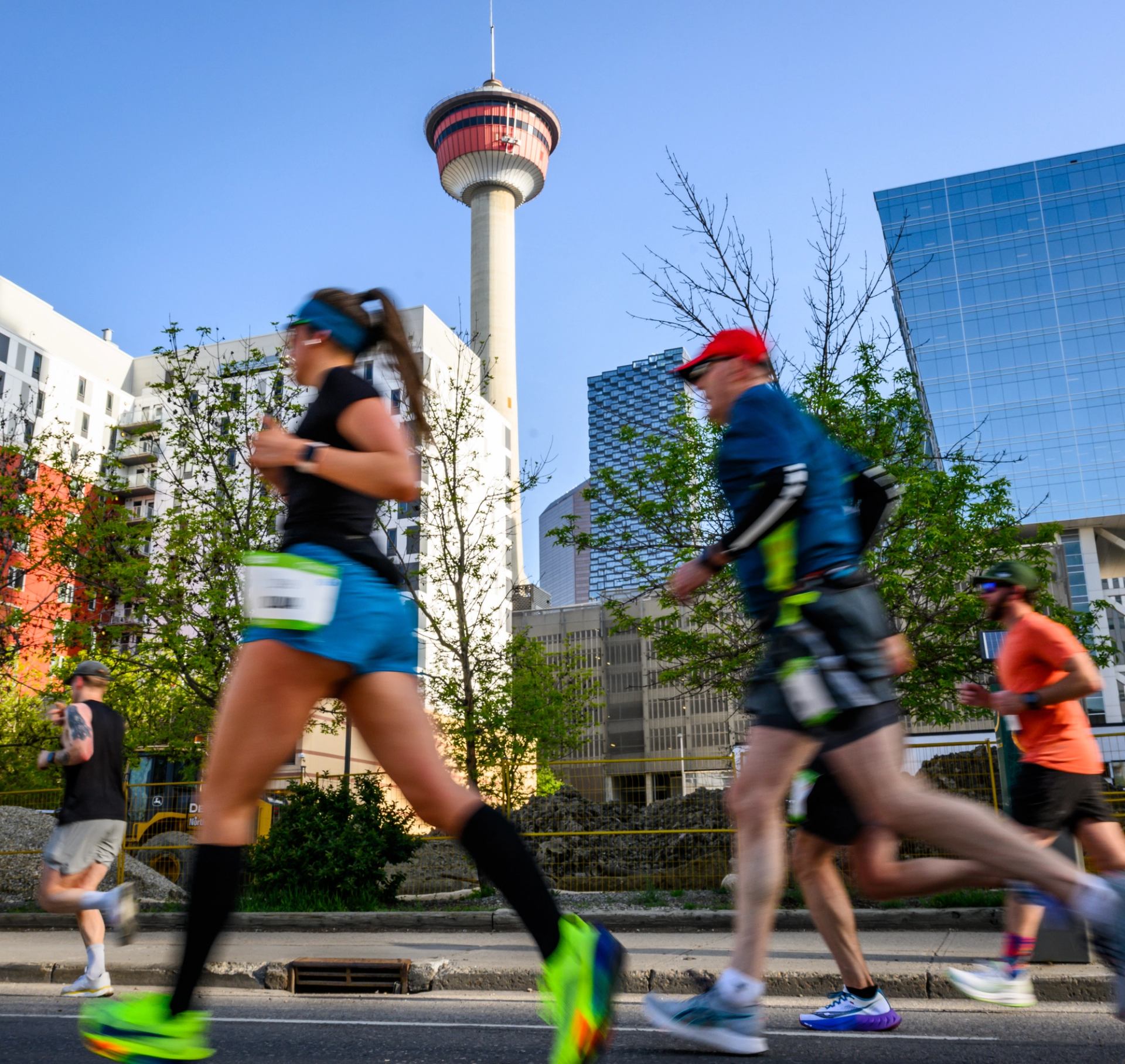 Runners pass Calgary Tower and downtown buildings along a city street.