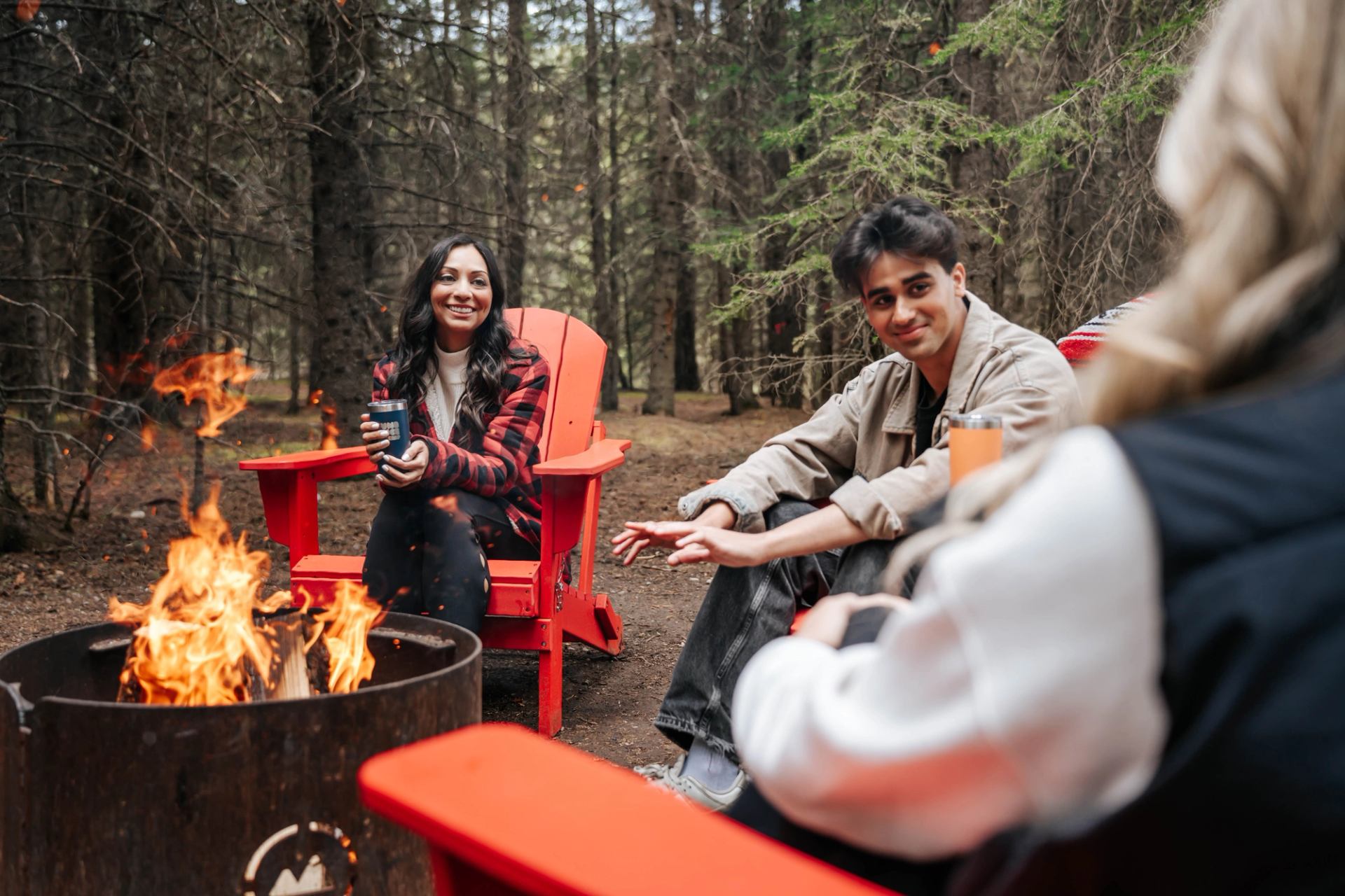People sit in red chairs around a campfire in a forest, holding insulated mugs.
