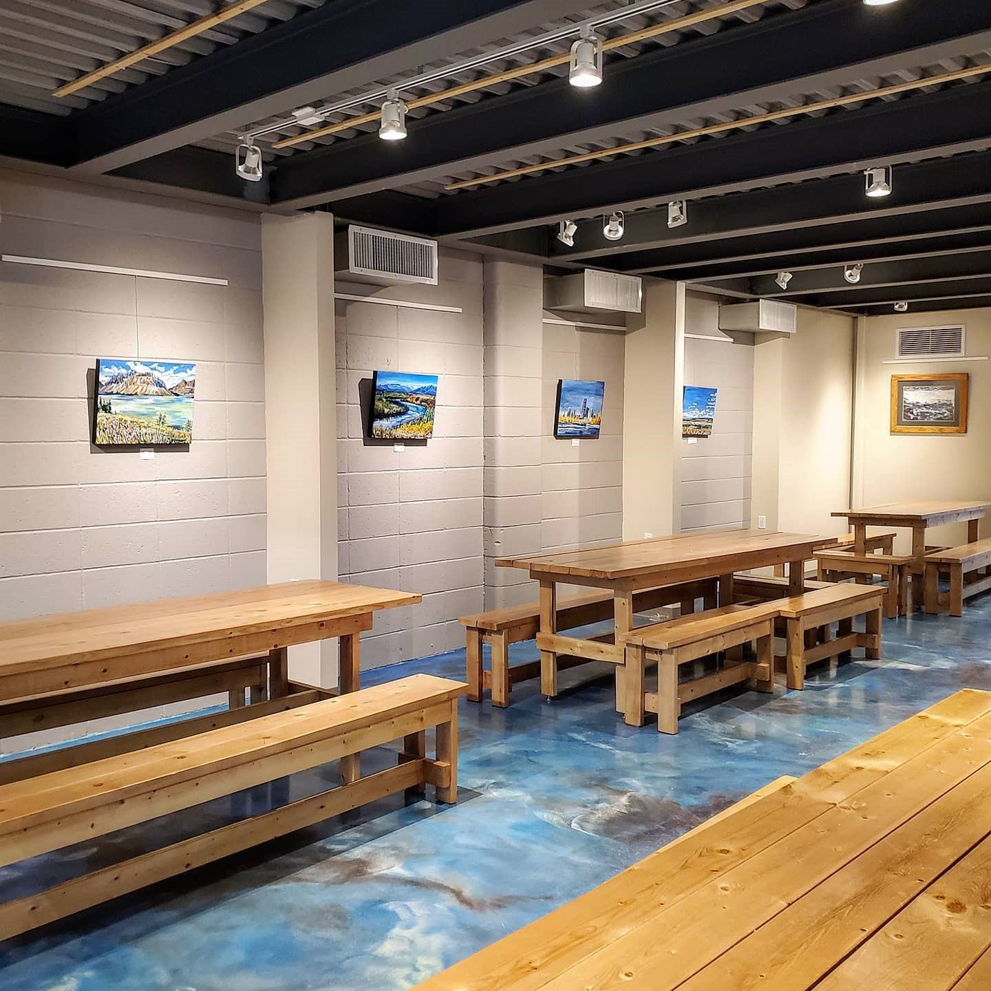Brewery seating area with long wooden tables, benches, and landscape paintings on walls.