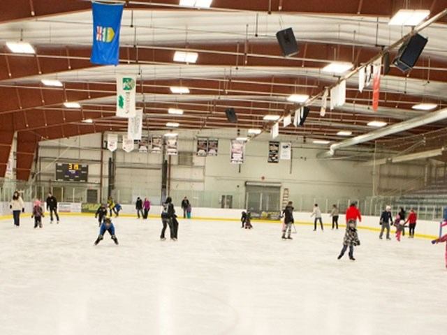 Indoor ice arena with hockey players on the rink and overhead scoreboards and lights.