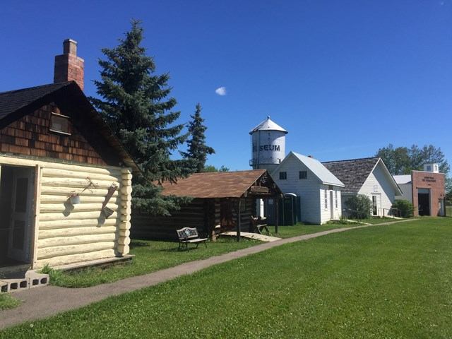 Historic village scene with log and wooden buildings under a clear blue sky. A museum water tower is visible. The mood is peaceful and nostalgic.
