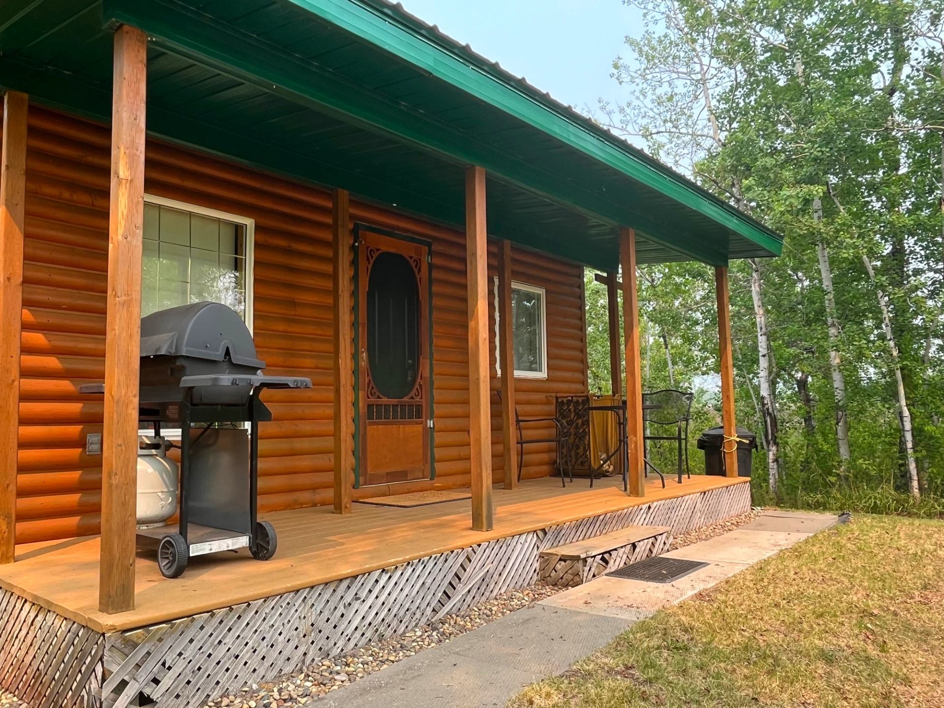 Wooden cabin with green roof, front porch, and barbecue grill surrounded by trees