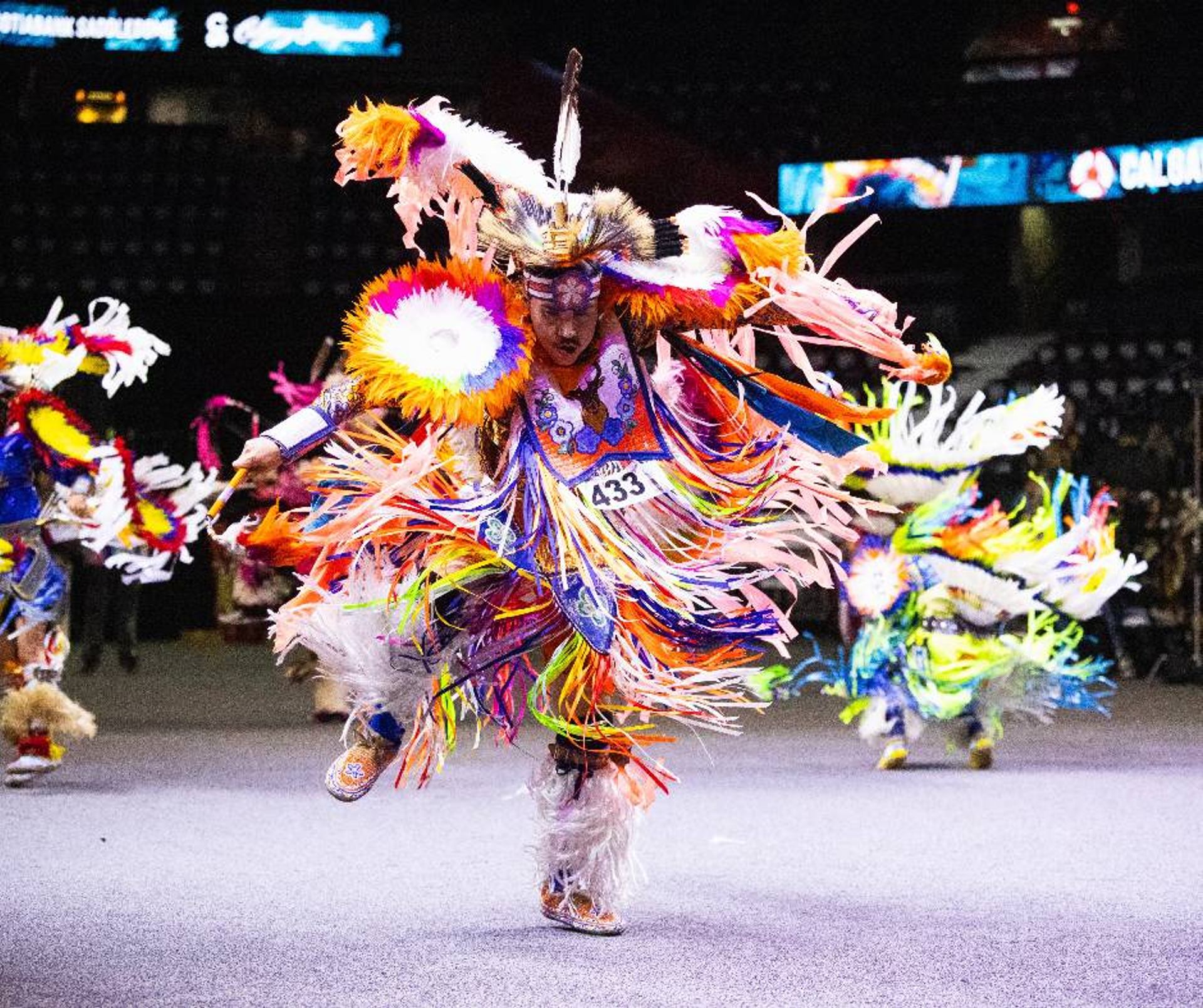 Powwow dancer in colourful regalia moves across arena floor during competition.