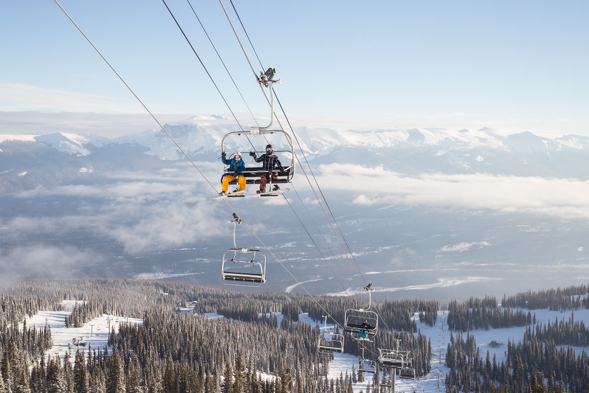 Skiers riding a chairlift above snowy slopes with mountains and clouds in the distance.