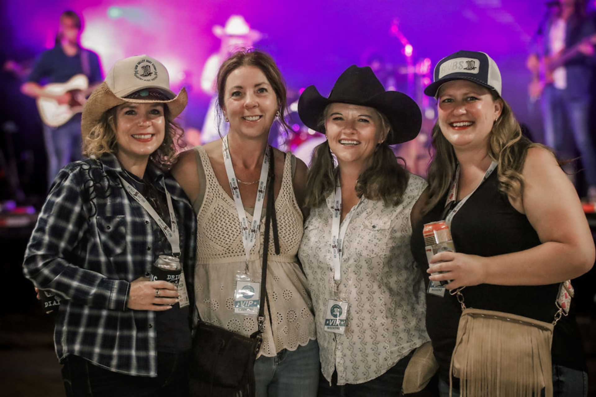 Four women smile in front of the stage. They have lanyards and drinks and hats. 