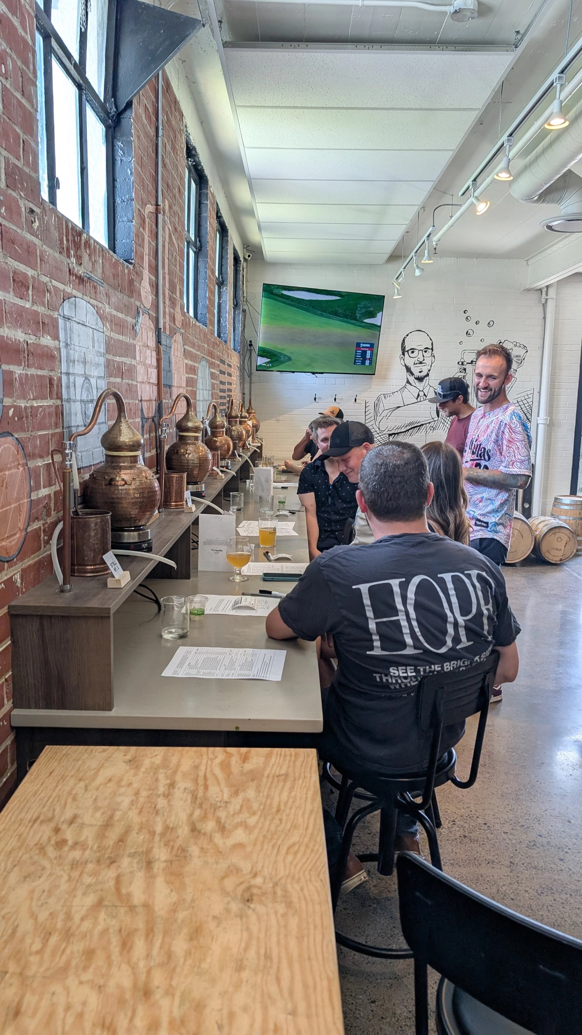 Guests sit at a tasting counter beside copper stills in a brick-walled distillery.