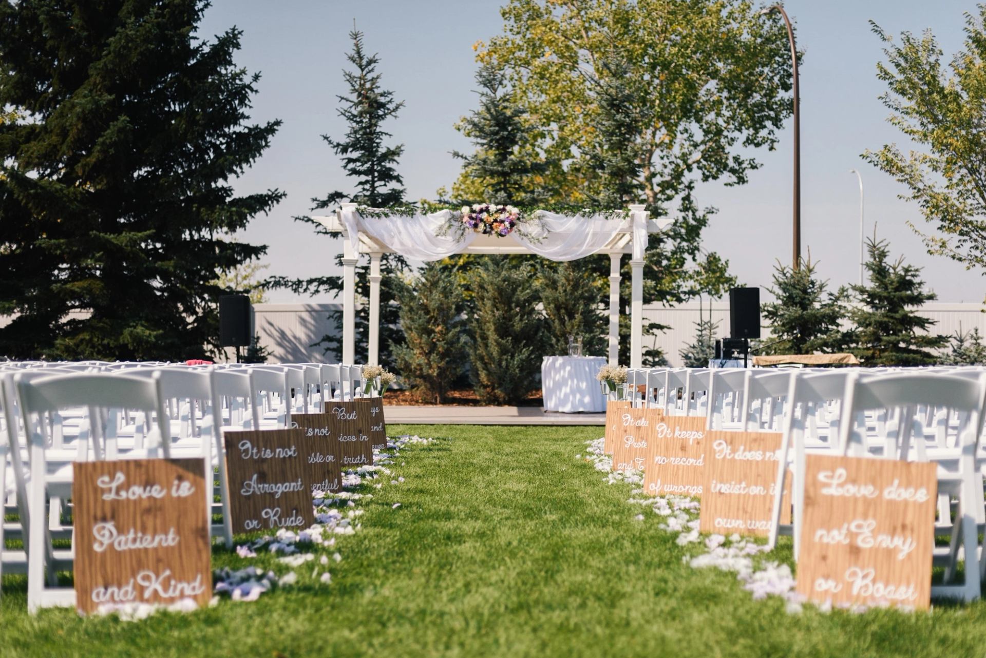 Outdoor ceremony aisle with white chairs, wooden signs and a flower-decorated arch.