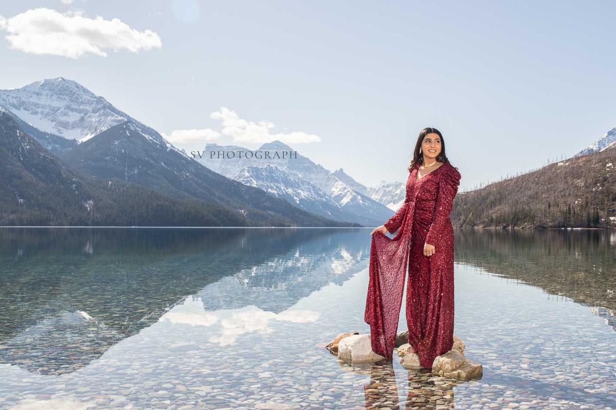 A person in a flowing red dress stands on stones in a clear lake with snowcapped mountains reflected.