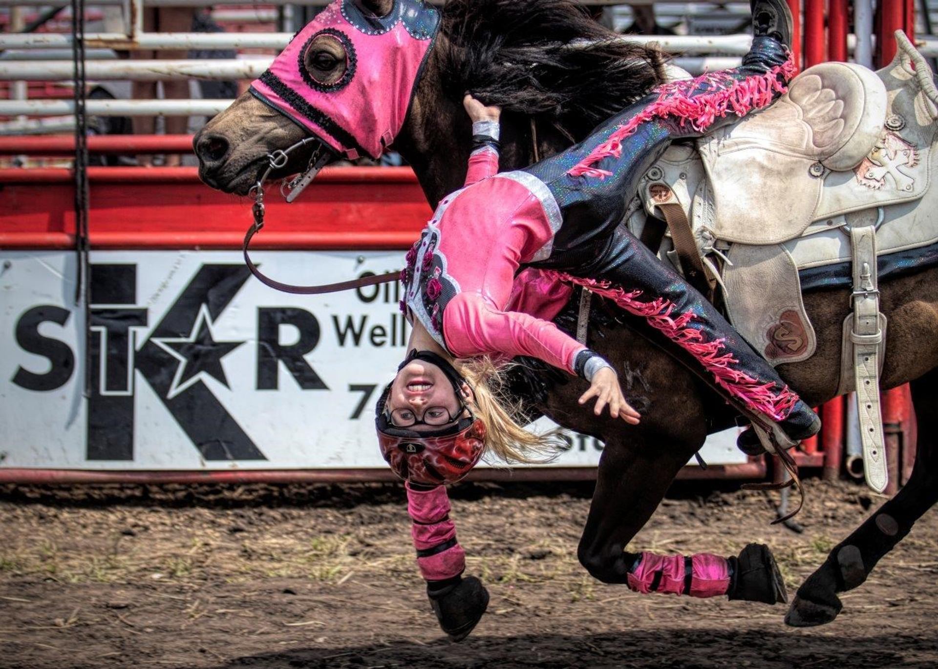 Trick rider hangs upside down from galloping horse in pink gear at rodeo arena.