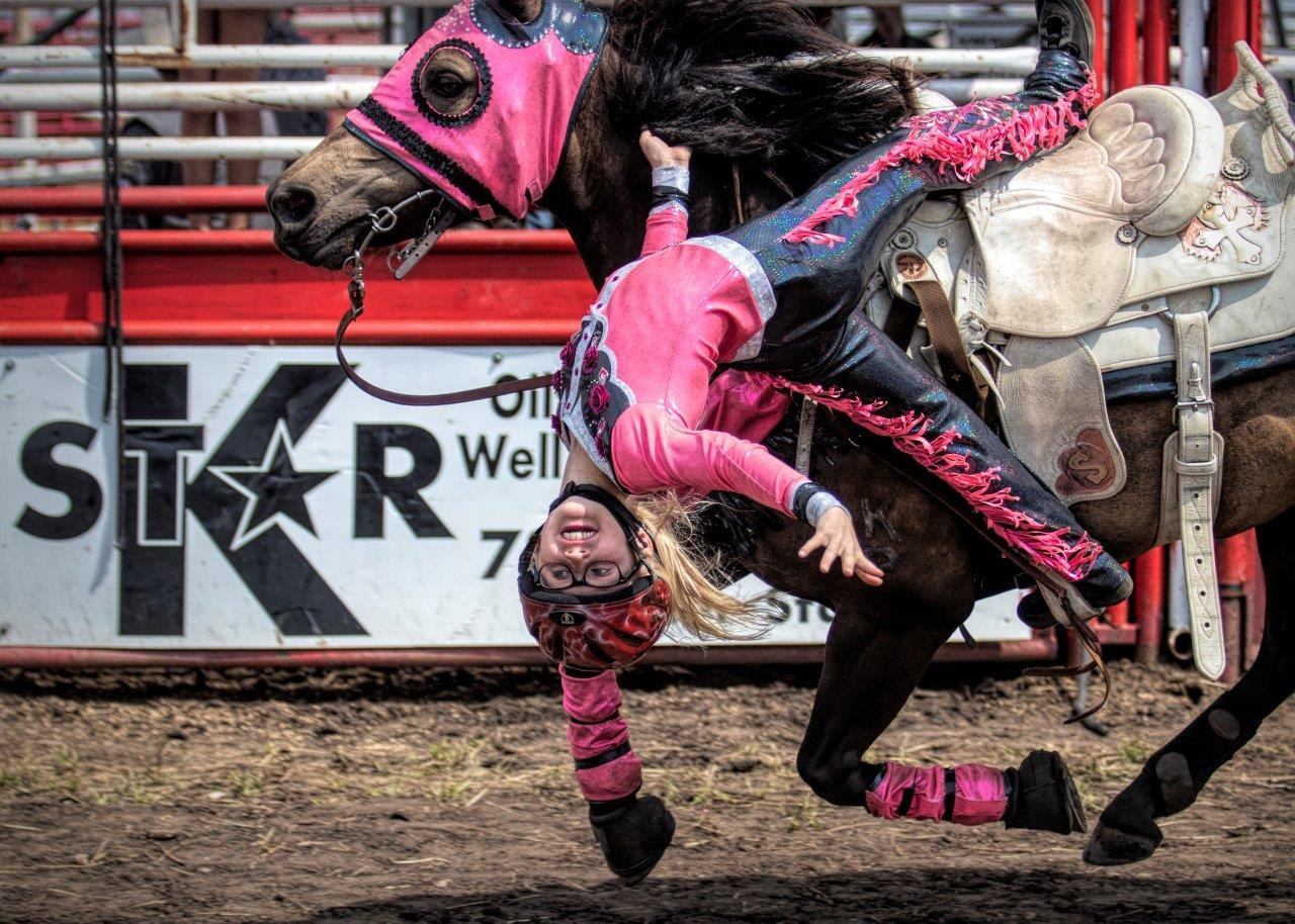 Trick rider hangs upside down from galloping horse in pink gear at rodeo arena.