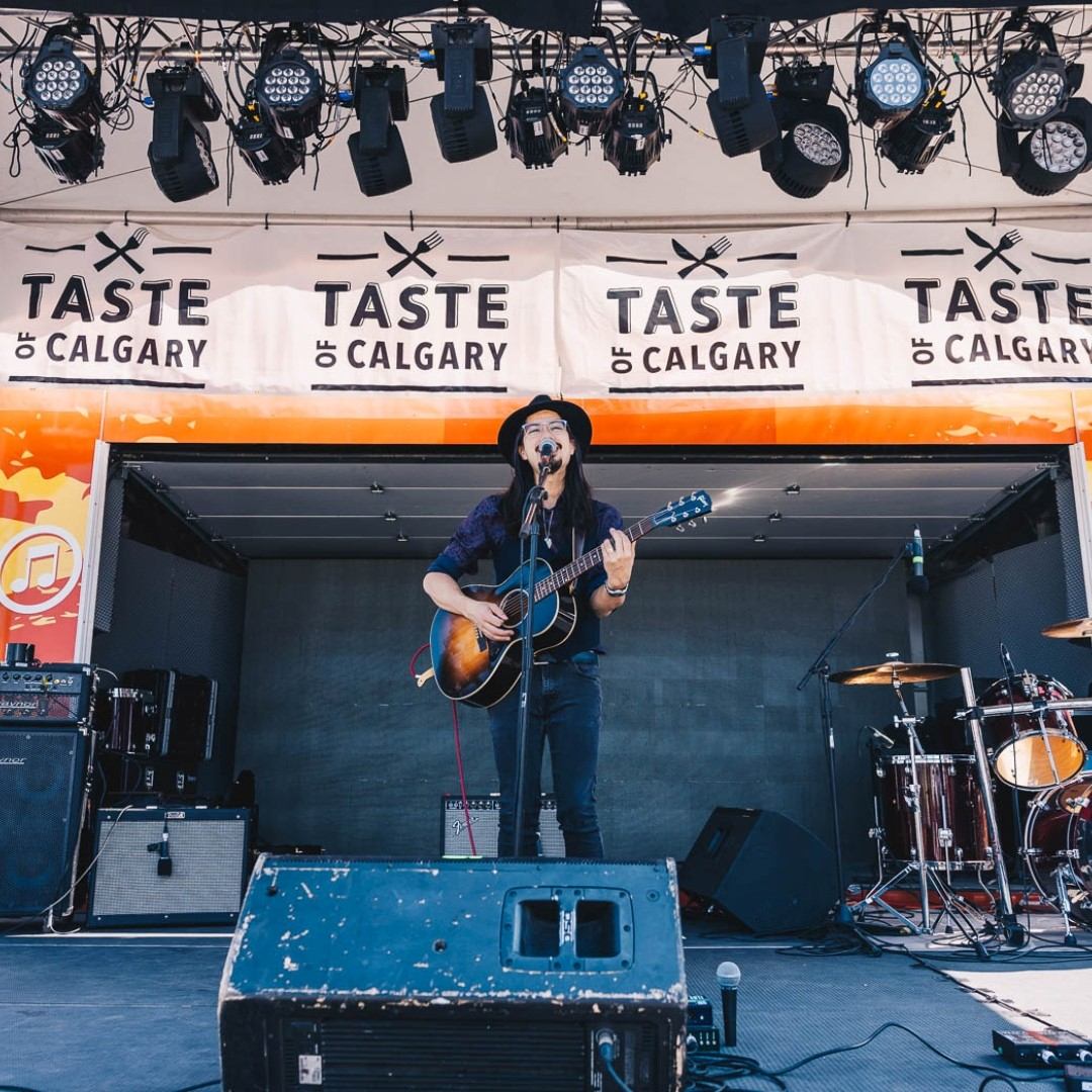 Musician plays guitar on stage under Taste of Calgary banner and lights.