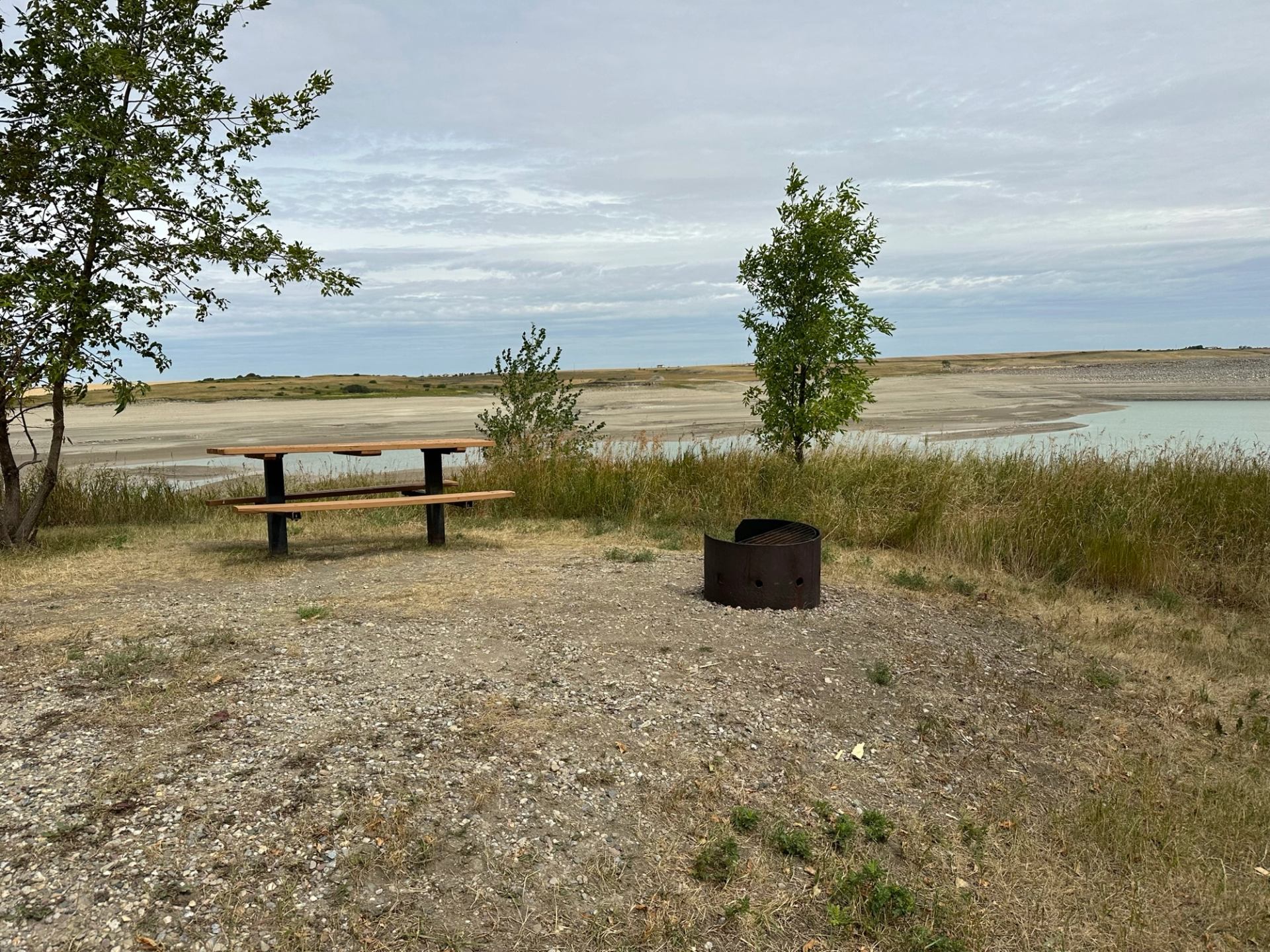 A picnic table and fire ring overlooking a calm shoreline and open prairie.