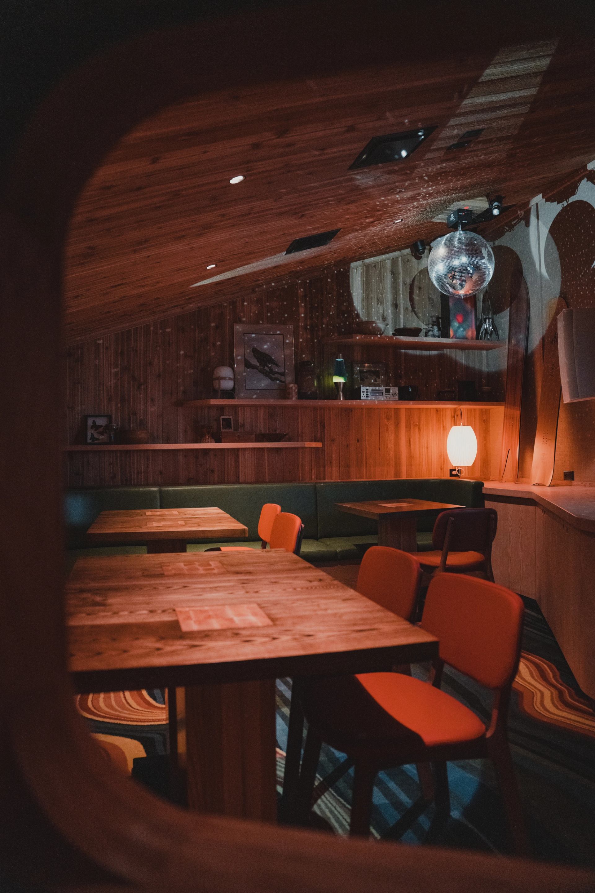 A warmly lit bar interior with wood paneling, orange chairs, green booths, a disco ball, and a wavy patterned floor, viewed through a circular frame.
