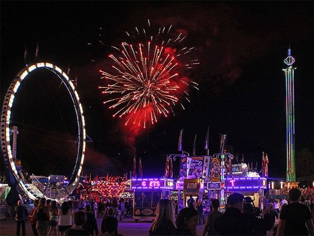 Fireworks bursting over a vibrant night carnival with bright rides and crowds.